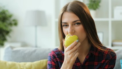 Beatiful brunette woman eating fresh green apple while resting on the sofa at home. Close up shot. - Powered by Adobe