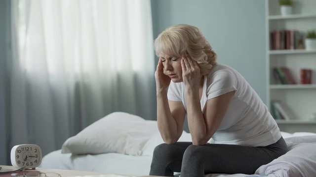 Senior woman sitting on bed and suffering from terrible headache, health problem