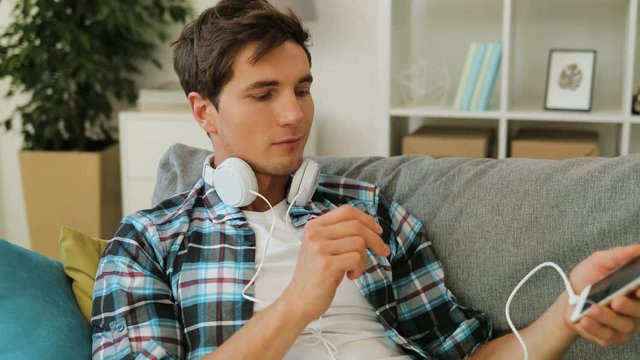 Attractive man wearing white headphones for listening to music on his smartphone while relaxing and lying on the sofa in bright living room.