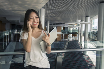 Adorable black-haired  Asian girl holding modern computer speaking on phone and cute smiling office area. Indoor portrait of young lady in white shirt calling boss carrying laptop to workplace.