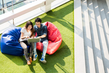 Asian team of man and women watch laptop computer while siting on bean bag. Study, relax, recreation concept. Colorful with copy space.