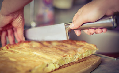 Cooking at home concept: vintage faded image of a woman’s hands cutting a potato pie with chef’s knife in the kitchen