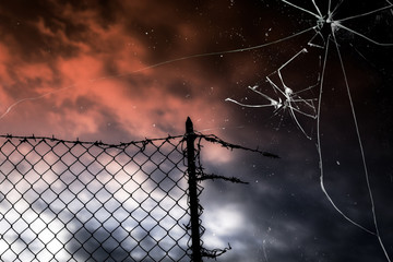 A broken fence over the sunset sky, barbed wire on top, seen through a broken glass. Symbolic shot:...