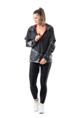 Fit active woman in legging zip tracksuit jacket fastener looking down. Full body length portrait...