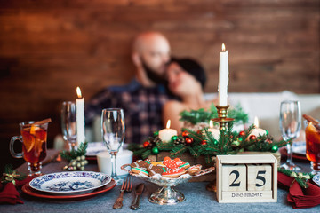 Beautiful young couple behind the decorated Christmas dinner table at the decorated New Year interior with tree. Christmas eve, calendar date of 25 on the table