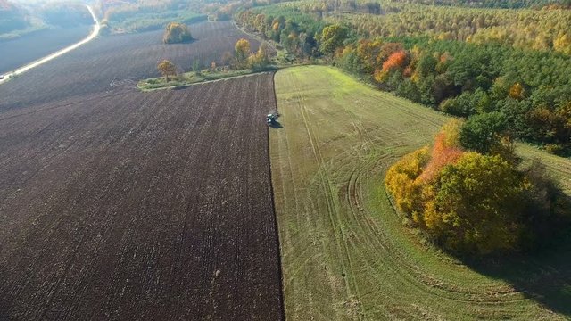 Aerial shot of tractor plowing black soil close to forest. Autumn sunset.