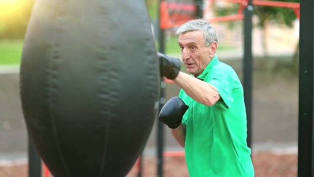 Boxer hits punching bag. Active elderly boxer in boxing glove training in a boxing club. Senior man boxing with a punching bag. Aged grey-haired man in green t-shirt training in outdoor gym