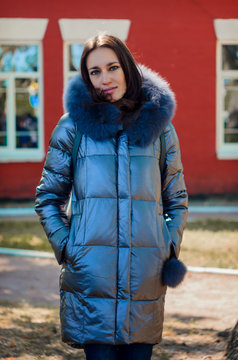 brunette woman stands under a tree in a park next to a red house in a winter jacket (down jacket)
