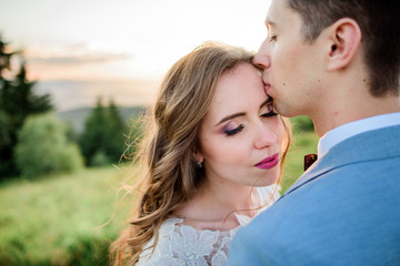 Groom kisses bride's forehead standing on a green hill