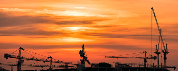The silhouette image of tower cranes at the real estate and building construction site with the background of golden sky of sun set.