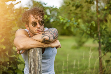 Portrait of handsome tattooed man in sunglasses leaning on pole looking away on natural background. 