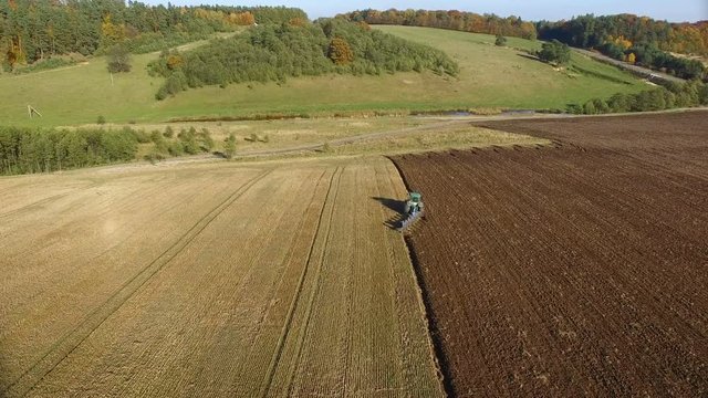 Aerial shot of tractor plowing black soil close to forest. Autumn sunset.