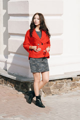 Young beautiful stylish woman dressed in a red coat, gray knitted dress, black boots. Street style, spring and autumn trend, flirty look, hair in the wind