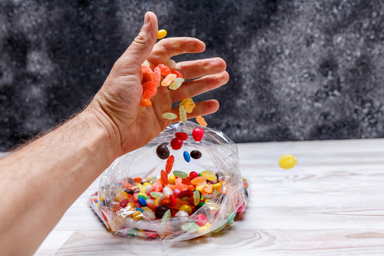 A man's hand pours out mix of colored candy in a plastic bag that lies on the light wooden surface of the table. The background is dark.