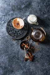 Glass cup of traditional indian masala chai tea with ingredients above. Cinnamon, cardamom, anise, sugar, black tea in glass teapot, jug of milk over dark texture background. Top view with space