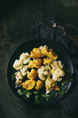 White and yellow cauliflower gratin served on black ceramic plate with coriander leaves, textile napkin and cutlery over dark metal texture background. Flat lay with space. Rustic style