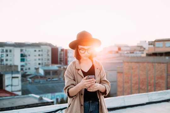 Amazing epic shot of young woman or teenager in coat and fedora hat standing on top of rooftop in big city, uses smartphone to chat or message application during sunset