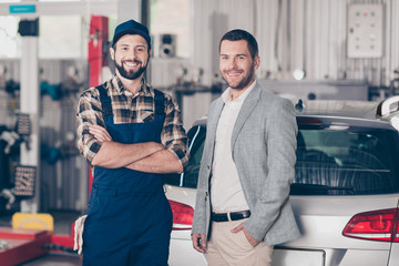 Closeup of two attractive guys, cheerful specialist repairman in blue overall, businessman in formal wear, man`s property silver car. Vehicle breakdown, check, inspection, engineering concept