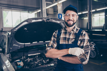 Attractive caucasian brunet bearded expert mechanic standing with metal mechanical keys in arm, in special safety outfit uniform, checkered shirt, hat head wear, open hood of vehicle