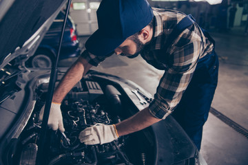 Fototapeta na wymiar Close up of young bearded expert specialist appraising the damage for repairing car at work shop, mounting it, arms in white knitted gloves, hat and special blue uniform overall, ckeckered shirt