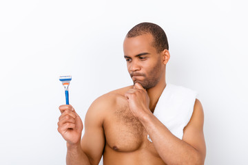 Fototapeta premium Skeptic, unsure, uncertain, doubts concept. Young african nude athletic guy is looking sceptical at razor, has a grimace of distrust on white background