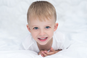 Closeup of smiling little boy lying on white blanket on the bed. Happy cute child with big blue green eyes looking at the camera