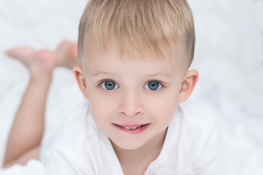 Closeup of smiling little boy lying on white blanket on the bed. Happy cute child with big blue green eyes looking at the camera