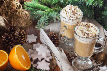 Christmas background with two latte, cookies star, fir branches, cones, orange and spices, close up. Couple coffee with milk and homemade tasty cookies in tray on wooden background. Top view.
