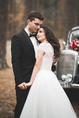 Naklejka premium Stylish wedding couple, bride, groom kissing and hugging near retro car in autumn