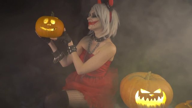 Sexy devil girl with gray hair and horns in red short dress, caresses kisses pumpkin. Celebrate halloween with scary funny pumpkins in smoke. Demon woman in purgatory. Gates of hell in fire