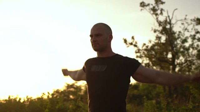 slow motion round picture of happy bald muscular man wearing black T-shirt warming up rotating arms enjoying time on fresh air on the edge of hill over sea in summer