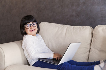 Smiling little girl with glasses using her laptop while sitting on the couch at home