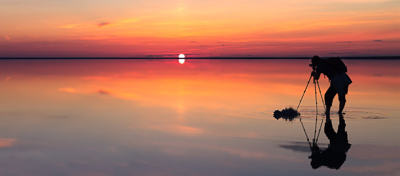 Silhouette of alone man takes a picture of a vibrant sunset reflected in shallow waters of solt lake. Banner size