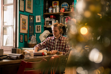 Fototapeta na wymiar Young adult man with long blonde hair wrapping christmas present with red ribbon seated on wooden table in cozy apartment indoor.