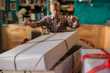 Young adult man with long blonde hair wrapping christmas present with red ribbon seated on wooden table in cozy apartment indoor.