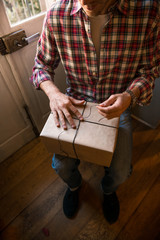 Young adult man with long blonde hair wrapping christmas present with twine ribbon seatednear windows in cozy apartment indoor.