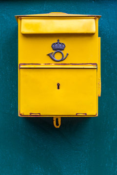 Close up of a classic yellow public postal mail box with a mail symbol, mounted on a blue wall outdoors.