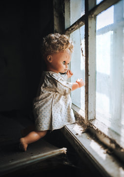 The old doll stands by the window and looks out the window. The concept of loneliness, sadness, poverty. Horror, scary, Halloween.