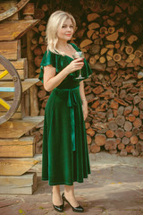 A middle-aged woman in a vintage green velvet dress and emerald accessories drinks red wine in the wood. The concept of vintage and retro femininity