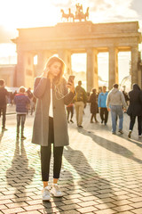 Full length portrait of young woman with earphones listening to music on phone while standing in front of Brandenburg gate in Berlin, Germany. © Bojan