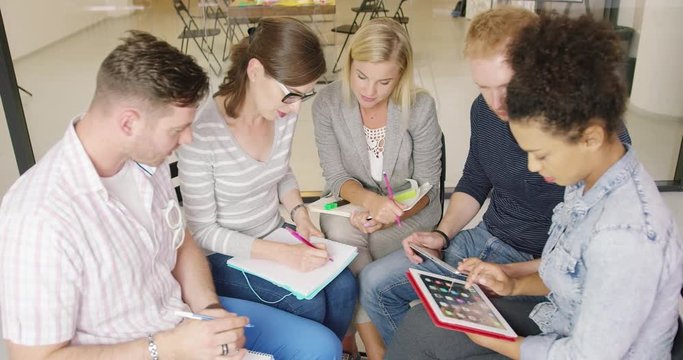 Group of colleagues in casual clothes sitting in office, discussing very important project and making notes in notebooks and on tablet.
