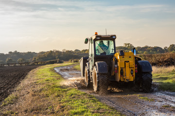 Obraz premium Tractor driving along a wet farm track with a ploughed field to the left.