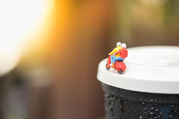 Travel Concept. Two traveler miniature figures ride motorcycle / scooter on cover of plastic cup take away of iced black coffee.