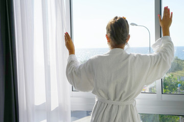 Young beautiful woman standing near the floor window, looking at the sea.  Sunny morning.