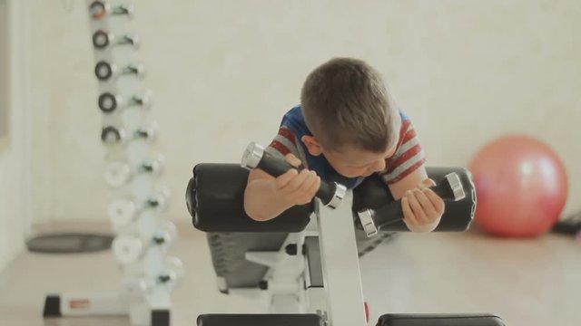 Smiling Young boy doing heavy dumbbell exercise for biceps. Baby working out with dumbbells on training apparatus. Strength and motivation. The concept of a healthy lifestyle. GYM
