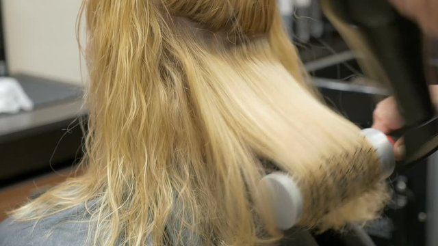 A stylist barber combs her hair to a blonde woman using a hairdryer.