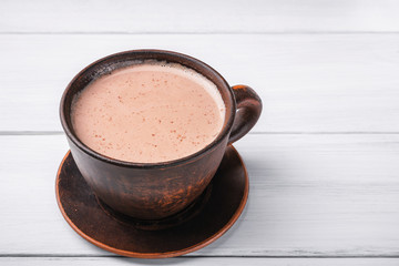 Hot cocoa with milk in brown clay cup, on table of white wooden planks