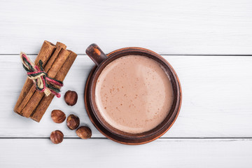 Heißer Kakao mit Milch in brauner Tontasse, Haselnuss- und Zimtstangen auf Tisch aus weißen Holzplanken, Ansicht von oben