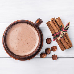Hot cocoa with milk in brown clay cup, hazelnut and cinnamon sticks on table of white wooden...