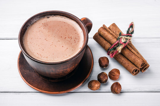 Hot cocoa with milk in brown clay cup, hazelnut and cinnamon sticks on table of white wooden planks